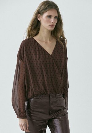 Donna che indossa una camicetta marrone trasparente a pois con maniche lunghe e pantaloni di pelle marrone scuro, in piedi davanti a uno sfondo neutro.