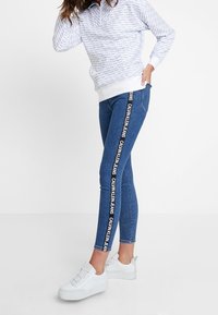 Jeans ajustados de denim azul oscuro con detalle del logo en el lateral, combinados con una sudadera ligera blanca con un estampado repetitivo. Zapatillas blancas.