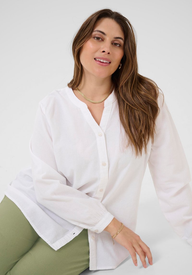 Femme avec de longs cheveux bruns portant une blouse blanche à boutons, un collier en or, un bracelet et un pantalon vert, posant devant un fond gris clair.