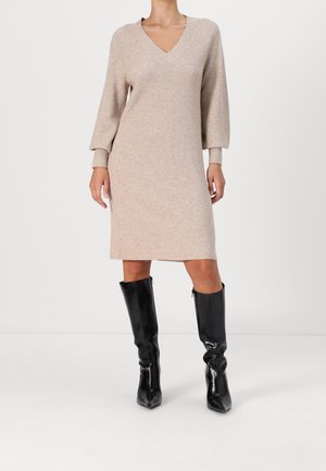 Robe en tricot beige à décolleté en V avec des manches longues, présentant une texture côtelée. Associée à des bottes noires brillantes jusqu'aux genoux.