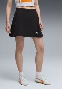 Jupe de sport noire plissée avec une taille ajustée et un logo Puma blanc. Associée à des baskets bleu clair avec des accents orange et des chaussettes blanches.