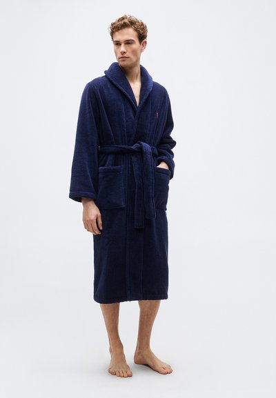 Άνδρας που στέκεται ξυπόλητος φορώντας μια μπλε βαμβακερή ρόμπα terry με γιακά κασκόλ, μακριά μανίκια, ζώνη και μπροστινές τσέπες.
