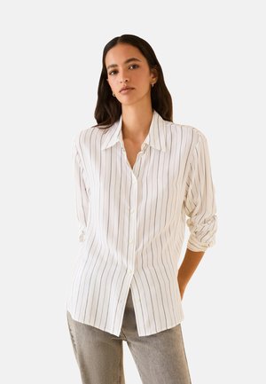 Donna con capelli lunghi e scuri che indossa una camicia bianca con sottili righe verticali nere e pantaloni grigi su uno sfondo uniforme.