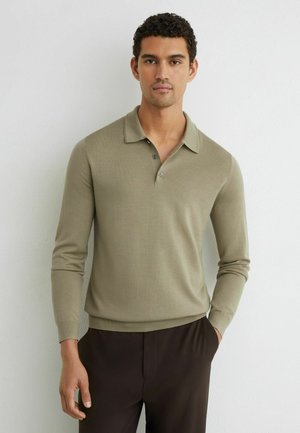 Man wearing a light olive green long-sleeve polo shirt and dark brown pants standing against a plain white wall.