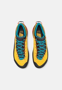 Scarpe da arrampicata con tomaia gialla, puntale nero, fodera turchese e lacci blu. Presentano dettagli rossi e una linguetta imbottita per il comfort.