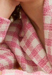 Růžová a bílá kostkovaná tkanina s texturovaným, tkaným vzorem. Tkanina vypadá silně a útulně, tvoří část límce nebo šály.