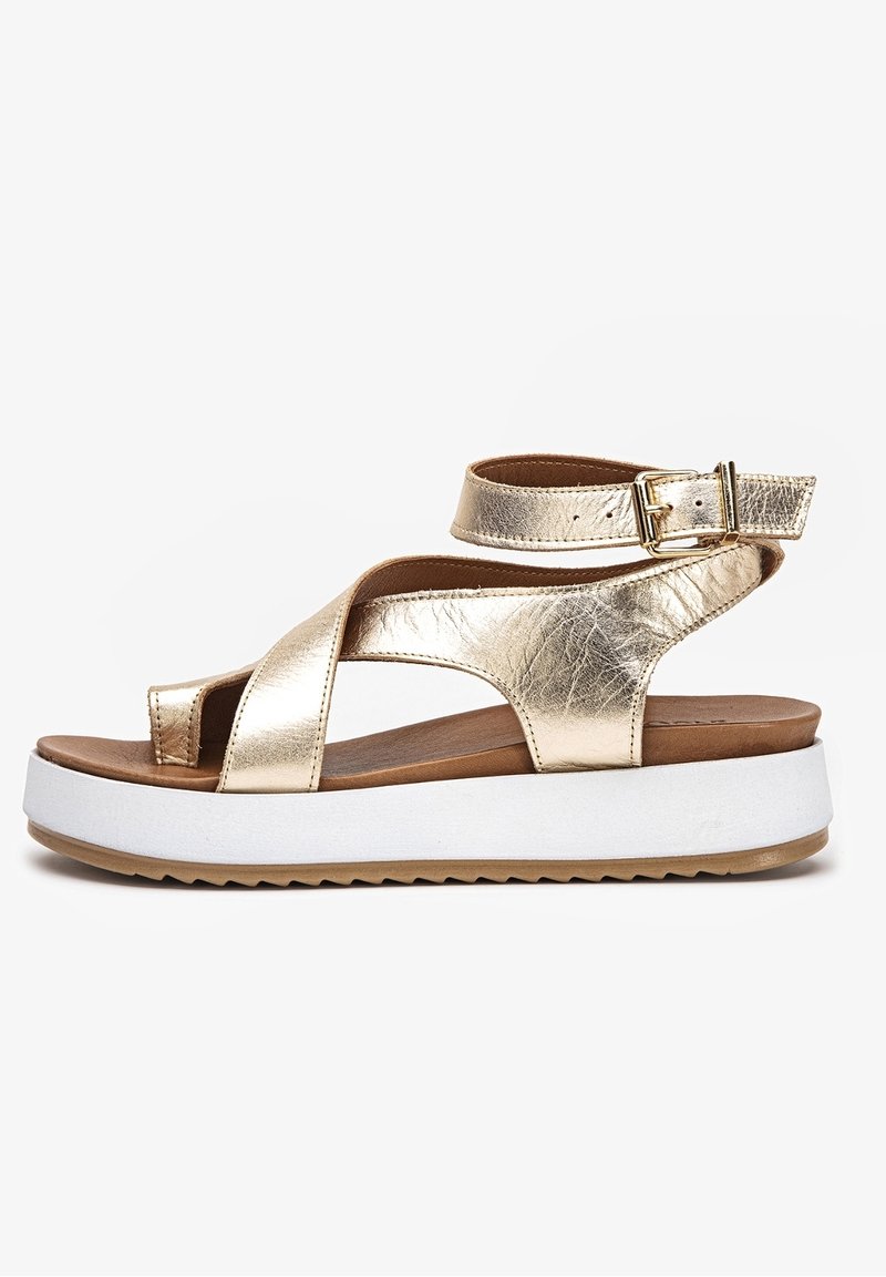 Złote metaliczne sandały z krzyżującymi się paskami, otwartymi palcami i paskiem na kostkę. Biała platforma z teksturowaną podeszwą dla lepszej przyczepności.