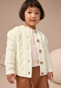 Cardigan en crème tricoté avec un motif en cable, boutons en bois, col rond et poignets côtelés, porté sur une chemise rose clair et un pantalon marron.