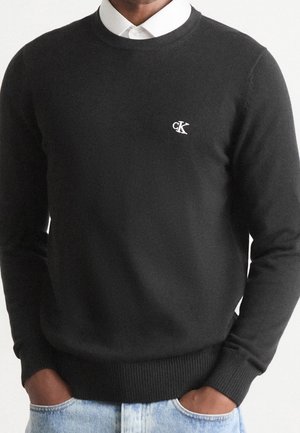 Mann trägt einen schwarzen Pullover mit Rundhalsausschnitt und weißem cK-Logo auf der Brust, darunter ein weißes Hemd mit Kragen, sowie hellblaue Jeans mit Händen in den Taschen.