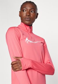 Dugi rukav sportski top u vibrantnoj ružičastoj boji. Sadrži visoki ovratnik sa zatvaračem i bijelim logotipom. Glatka, rastezljiva tkanina s otvorima za palčeve.