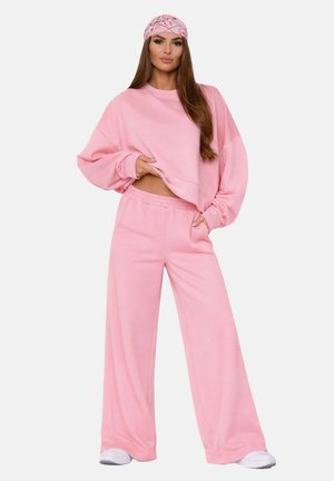 Femme portant un sweat-shirt rose ample, un pantalon rose à jambes larges, des baskets blanches et un foulard rose à motifs, debout avec une main dans la poche.