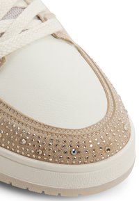 Sneaker med en beige ruskindsoverdel, hvid læder tåboks, struktureret cremefarvet mellemsål og prydet med små, blanke rhinsten langs kanten.