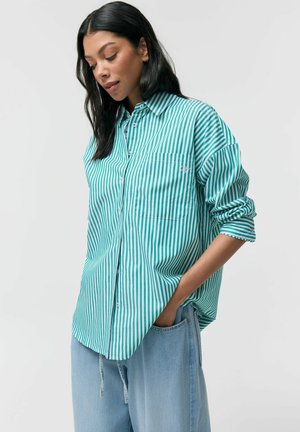 Kobieta o długich ciemnych włosach, ubrana w zieloną i białą koszulę z pionowymi paskami i jasnoniebieskie dżinsy, z jedną ręką w kieszeni.