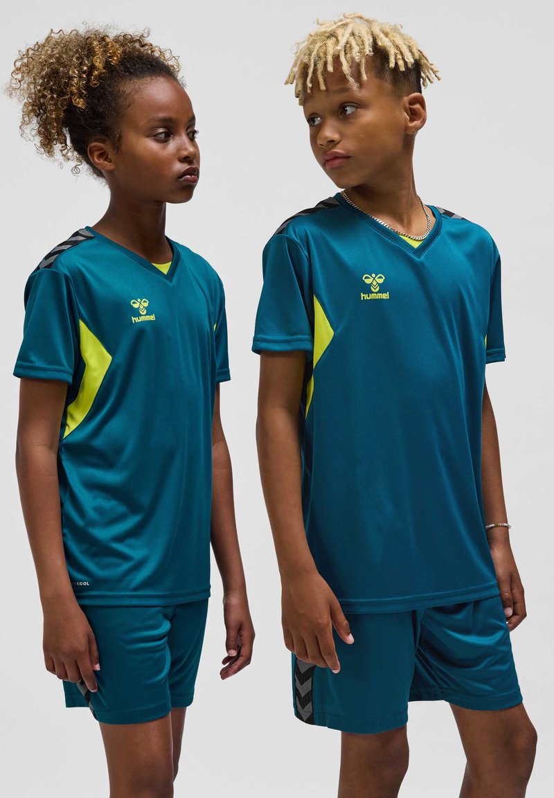 Deux enfants en tenue de sport bleu sarcelle avec des accents jaunes, debout et se regardant sur un fond uni.