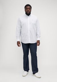 White long-sleeve button-up shirt with small blue polka dots, paired with dark blue jeans and white sneakers, standing on a neutral background.