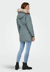 Parka grigia imbottita con cappuccio foderato in pelliccia, vita elastica e tasche laterali, abbinata a jeans skinny blu chiaro e stivali neri alla caviglia.