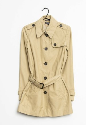 Trench beige avec des boutons noirs et une ceinture à la taille, suspendu sur un cintre noir sur fond blanc.