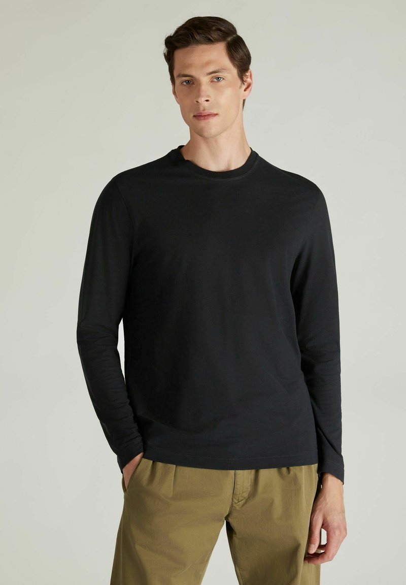 Schwarzes langärmliges T-Shirt aus einem glatten, leichten Stoff mit Rundhalsausschnitt, kombiniert mit Khaki-Hosen und Hände in den Taschen.