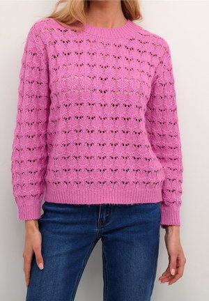 Frau trägt einen rosa Strickpullover mit einem strukturierten Lochmuster, kombiniert mit blauen Jeans, stehend vor einem schlichten weißen Hintergrund.