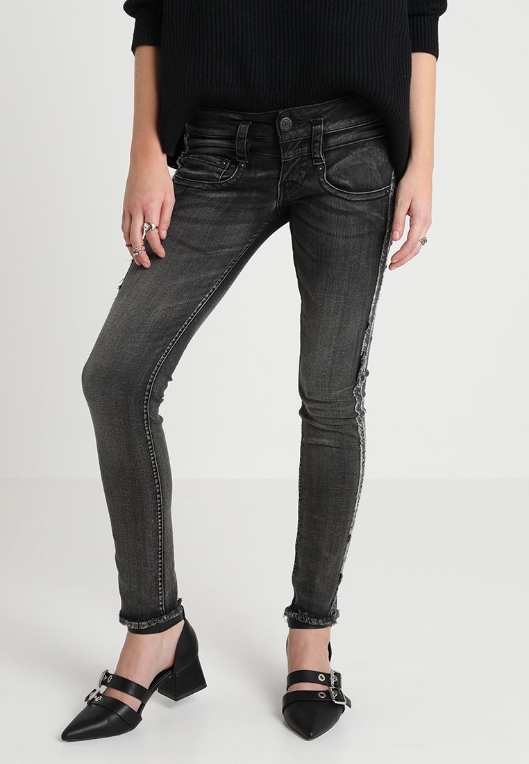 Jeans ajustados negros con acabado desgastado, dobladillos deshilachados y detalles de costura plateada, combinados con zapatos negros de tacón puntiagudos.