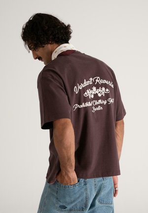 Man with curly hair wearing a brown T-shirt with white floral text on back and light blue denim shorts, hand in pocket, facing away.
