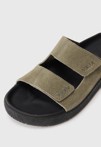 Grønn semsket sandal med to brede borrelåsremmer, svart gummisåle, teksturert overflate og preget logo-detalj på siden.