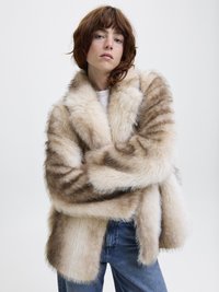Manteau en fausse fourrure beige avec de subtiles rayures brunes, doté d'un large col et d'une coupe ample. Assorti à un jean en denim. Fond uni.