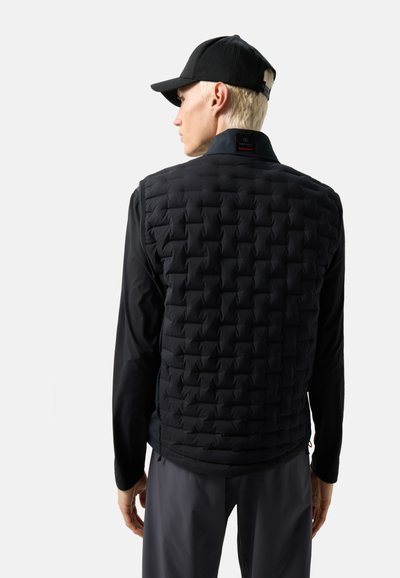 Mann mit kurzen blonden Haaren trägt eine schwarze gesteppte Weste, ein schwarzes Langarmshirt, graue Hosen und eine schwarze Baseballkappe, von hinten gesehen.