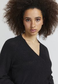 Melnā adītā džemperī ir V veida kakla izgriezums un teksturēta auduma materiāls, ar garām piedurknēm un brīvu piegriezumu.