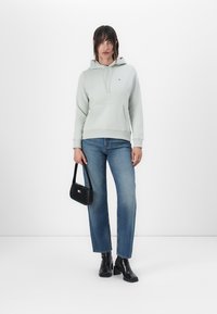 Hellgrünes Hoodie mit einer Vordertasche, kombiniert mit blauen Jeans. Schwarze Ankle Boots und eine kleine schwarze Tasche mit einem Logo-Akzent.
