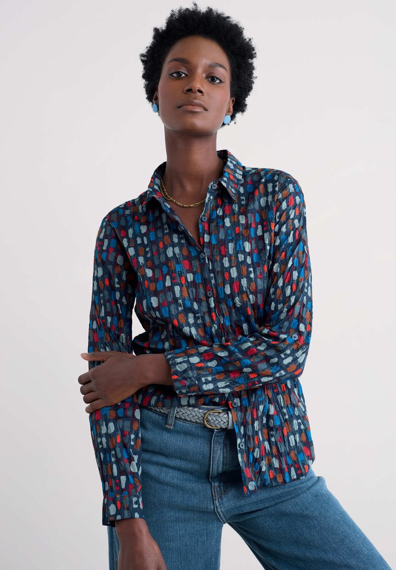 Mehrfarbige Bluse mit Knopfdesign, mit einem blauen Hintergrund und abstrakten Mustern in Rot, Orange und Weiß. Lange Ärmel.