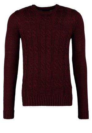 Mann geht, trägt einen dunkelroten Zopfstrickpullover, blaue Jeans mit hochgekrempelten Säumen, schwarze Stiefel und hält eine schwarze Tragetasche.