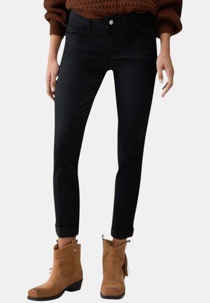 Persona che indossa jeans neri skinny risvoltati, maglione marrone a trama lavorata e stivaletti alla caviglia in suede color cognac con tacco basso e dettagli con nappine.