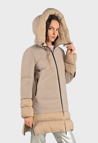 Beige pufferjas met een grote capuchon, contrasterend onderste gedeelte, ritssluiting en zwarte accenten. Bevat zijzakken en heeft een gemiddelde lengte.