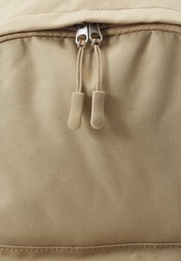 Mochila de tela beige con una superficie texturizada, que presenta un cierre con cremallera y dos tiradores tipo toggle. La costura a lo largo del borde superior es visible.