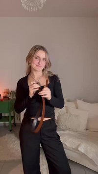 Haut noir à manches longues et pantalon noir à jambes larges. Ceinture en cuir marron clair tenue à la main. Fond neutre avec des accessoires doux.