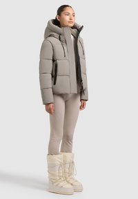 Veste grise rembourrée avec capuche, présentant des accents de zip noirs et des poches. Associée à des leggings beiges et des bottes blanches imperméables.