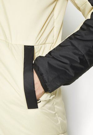Hand in beigefarbener Jackentasche mit schwarzem Besatz, trägt schwarzen Ärmel, Nahaufnahme zeigt Stoffstruktur und Nähdetails.