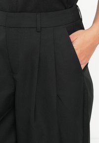 Pantaloni neri su misura realizzati in tessuto testurizzato, con pieghe frontali e tasche laterali, accompagnati da una vita liscia.