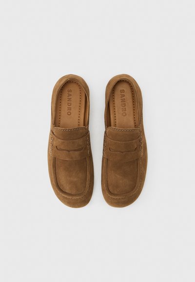 Ζευγάρι καφέ μοκασίνια από σουέντ με ραφές και λουράκι στο πάνω μέρος, με την επωνυμία "SANDRO" στην εσωτερική σόλα, σε ανοιχτό φόντο.