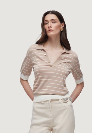 Kobieta z długimi ciemnymi włosami, ubrana w krótkim rękawem polo w beżowe i białe paski oraz kremowe spodnie z ozdobnymi kieszeniami.