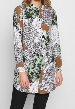 Femme portant un chemisier à manches longues boutonné avec des motifs floraux et abstraits verts, blancs, marron et noirs, associé à un pantalon noir.