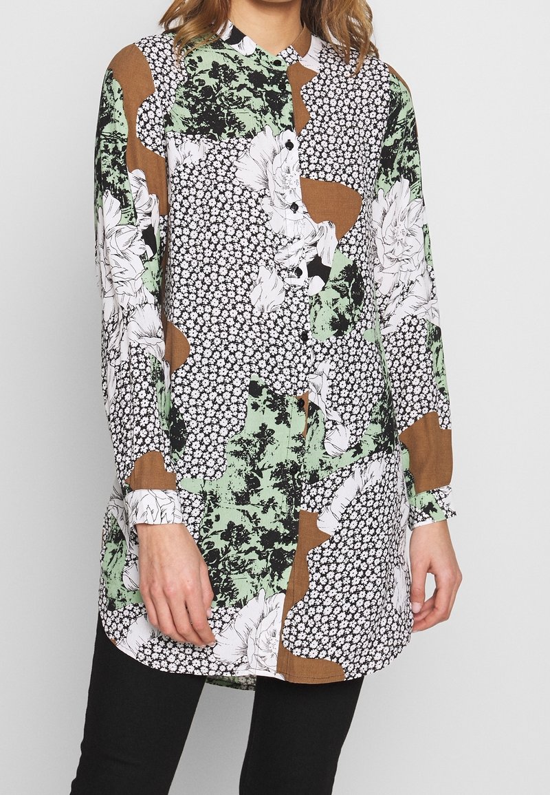 Femme portant un chemisier à manches longues boutonné avec des motifs floraux et abstraits verts, blancs, marron et noirs, associé à un pantalon noir.
