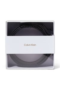 Zwarte leren riem gewikkeld in doorzichtig verpakkingsmateriaal, met een witte band waarop "Calvin Klein" in zwarte tekst en gouden accenten staat.