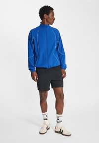 Blå lättviktjacka med vita linjedetaljer, dragkedjestängning och elastiska ärmslut, tillsammans med svarta shorts och vita, mönstrade strumpor.