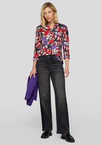 Bluse mit Blumenmuster in Rot und Lila mit drei Viertel-Ärmeln, kombiniert mit dunklen weit geschnittenen Jeans und einem gefalteten lila Pullover, der über dem Arm hängt.