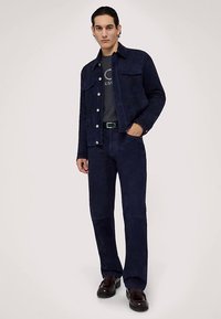 Marinblå mockajacka med två bröstfickor, kombinerad med matchande marinblå byxor. Modellen bär en grå t-shirt och mörka skor.