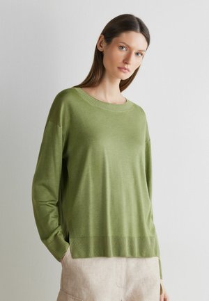 Kvinde med glat brunt hår, der har en løstsiddende lysegrøn sweater og beige bukser på, står foran en ensartet lyse baggrund.