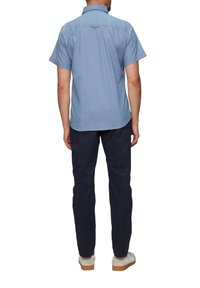 Himmelblaues Kurzarmhemd mit abgerundetem Saum, Schulterpasse und einer Schlaufe auf der Rückseite, kombiniert mit dunklen Hosen und hellen Schuhen.