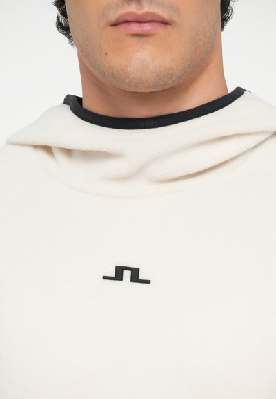 Cremfarbener Hoodie mit schwarzem Kragen und Logo. Weiches, strukturiertes Material und eine große, hängende Kapuze. Minimalistisches Design ohne zusätzliche Muster.
